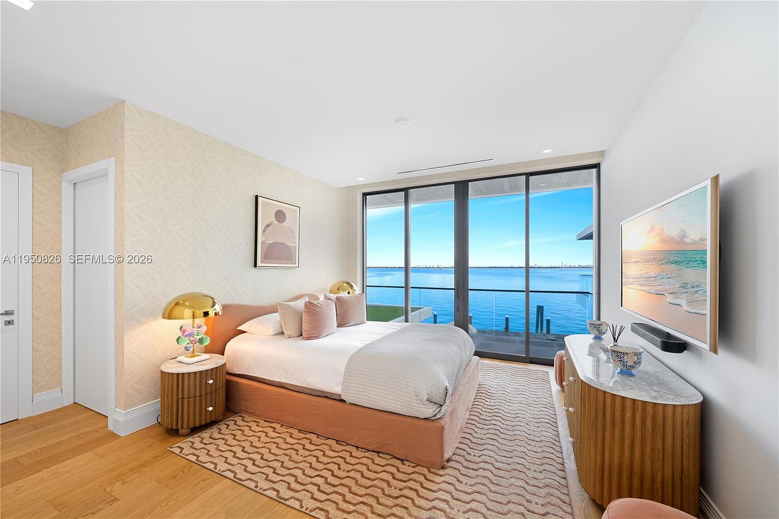 This is a primary bedroom featuring a large bed with a pink frame and neutral bedding, flanked by a wooden nightstand with a gold lamp. A large window offers a stunning view of the water, and a modern dresser with a marble top holds decorative items and a television. The room is decorated in neutral tones with a patterned rug, creating a serene and luxurious atmosphere.