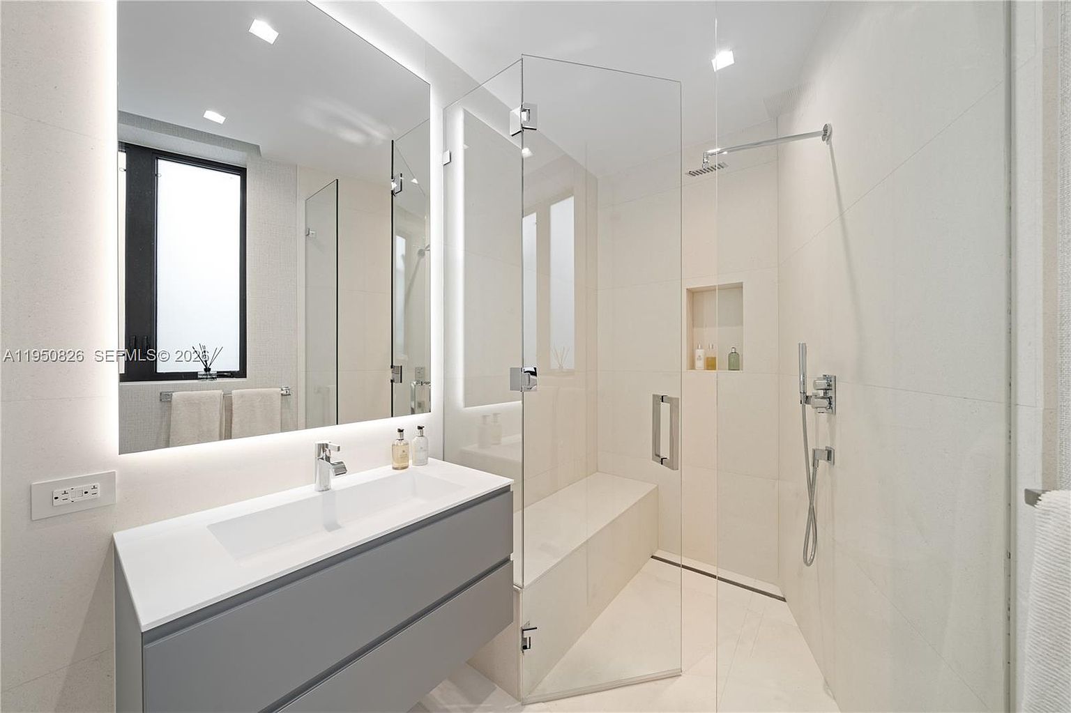 This is a modern primary bathroom featuring a sleek vanity with a gray cabinet and white countertop, a large mirror with integrated lighting, and a glass-enclosed shower with a built-in bench and niche. The bathroom is finished with light-colored tiles, creating a bright and clean aesthetic. The overall impression is luxurious and contemporary.
