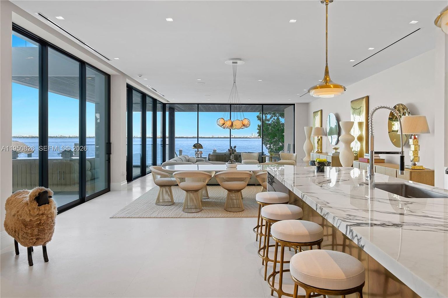 This is a bright and airy dining area with floor-to-ceiling windows offering stunning water views. The space features a modern dining table with unique chairs, a stylish light fixture, and a marble countertop island with bar stools. The overall design is contemporary and luxurious, emphasizing natural light and open space.