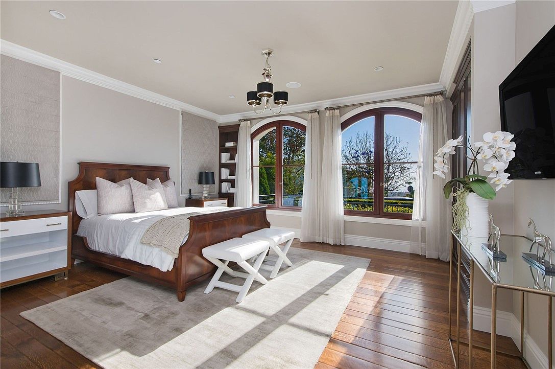 This elegant primary bedroom features a rich wood sleigh bed as the focal point, complemented by a neutral color palette and warm wood flooring. Large arched windows allow abundant natural light to fill the space, while a modern chandelier adds a touch of sophistication. The room is furnished with a sleek white dresser, a mirrored console table, and two white benches at the foot of the bed, creating a refined and inviting atmosphere.