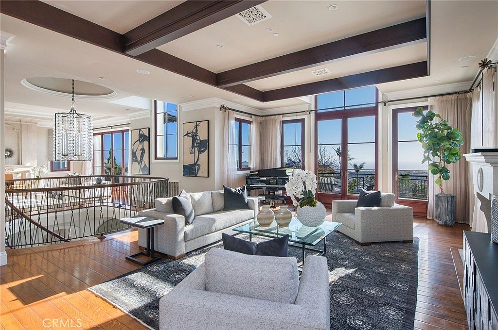 This expansive living room features a coffered ceiling with dark wood beams and a large, dark-toned area rug anchoring the seating area. The space is furnished with a light-colored sofa and matching armchairs, centered around a glass-topped coffee table, with a grand piano positioned near the tall windows that offer a view of the outdoors. A decorative metal railing overlooks a lower level, and a large, ornate chandelier hangs above the staircase area, contributing to the room's sophisticated and airy atmosphere.