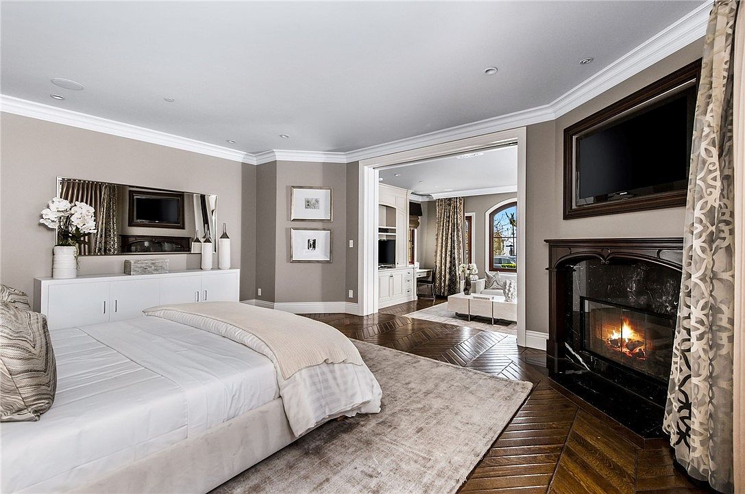 This elegant primary bedroom features a sophisticated neutral color palette, dark herringbone-patterned wood flooring, and a large area rug. A prominent fireplace with a dark stone surround and a mounted television serves as a focal point, while an open doorway leads into an adjacent sitting area. The room is well-lit with recessed lighting and includes a sleek white dresser topped with decorative vases and a mirror.