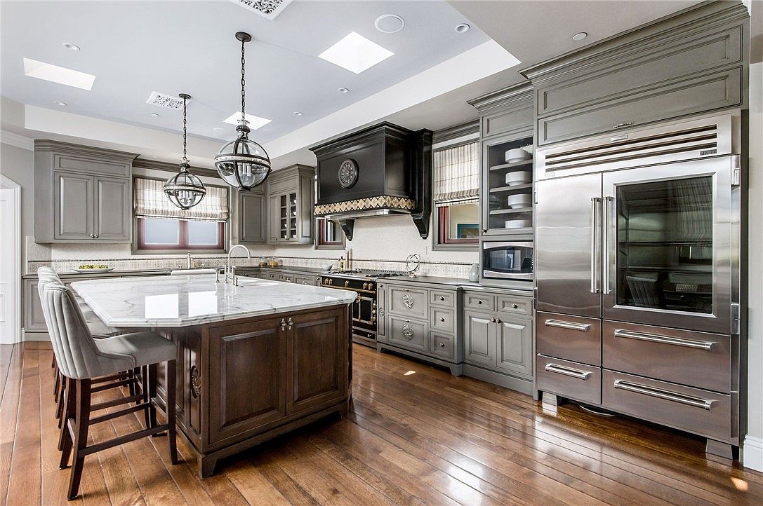 This spacious, high-end kitchen features a large central island with a stone countertop and bar seating, complemented by dark wood cabinetry and grey-toned lower cabinets. A prominent black range hood serves as a focal point above the stove, while stainless steel appliances, including a large double-door refrigerator, add a professional touch. The room is illuminated by decorative pendant lights and recessed lighting, with warm-toned wood flooring running throughout the space.