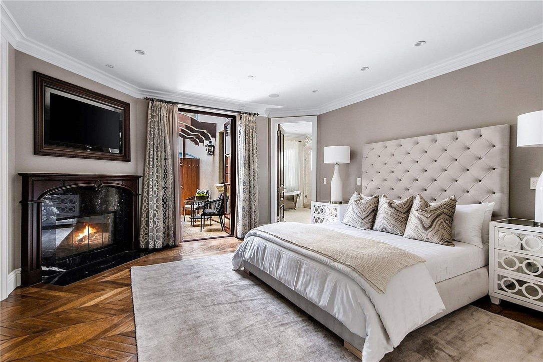 This elegant primary bedroom features a large, tufted upholstered headboard as the focal point, complemented by a neutral color palette and sophisticated decor. A dark wood fireplace with a stone surround adds warmth to the room, while a set of glass doors opens to a private outdoor patio. The space is finished with rich, dark wood flooring arranged in a herringbone pattern and a large area rug, creating a luxurious and inviting atmosphere.