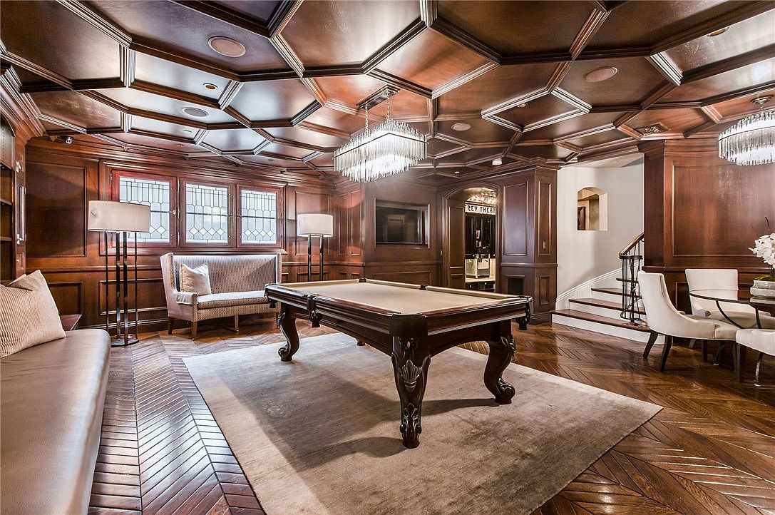 This sophisticated game room features a prominent pool table centered on a large area rug, set against a backdrop of rich, dark wood-paneled walls and a coffered ceiling. The space is illuminated by a modern crystal chandelier and tall, multi-paned windows that provide a classic, refined aesthetic. A leather sofa and a patterned settee offer comfortable seating, while a staircase in the background leads to another level, enhancing the room's sense of depth and luxury.