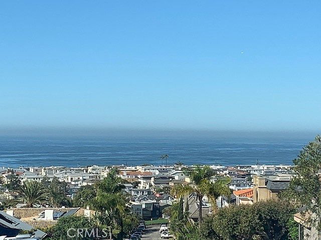 This elevated, wide-angle view captures a sprawling coastal neighborhood overlooking the vast blue ocean under a clear sky. The perspective highlights the dense residential layout, featuring various rooftops and lush greenery interspersed throughout the community. The scene emphasizes the property's prime location with expansive, unobstructed horizon views.
