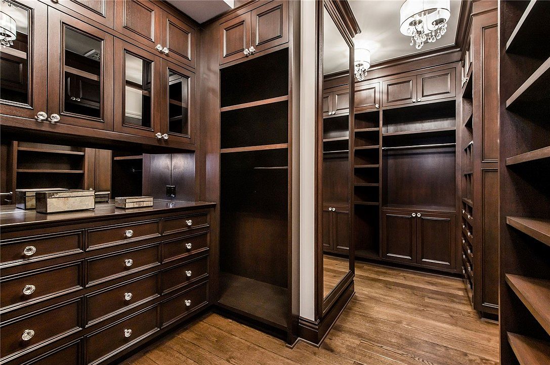 This luxurious walk-in closet features rich, dark-toned custom cabinetry with extensive shelving, hanging space, and multiple drawers. A full-length mirror is integrated into the cabinetry design, reflecting the warm lighting and the polished wood flooring. The space is organized and elegant, offering a sophisticated storage solution with a high-end aesthetic.