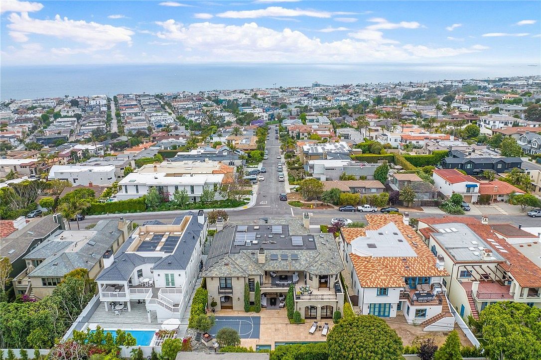 This high-angle aerial view captures a coastal residential neighborhood, focusing on a large, multi-story estate with a private pool and a basketball court in the backyard. The property features a complex roofline with solar panels, while the surrounding area showcases a dense collection of homes leading toward the ocean horizon. The perspective provides a comprehensive look at the property's layout, outdoor amenities, and its prime location within the community.