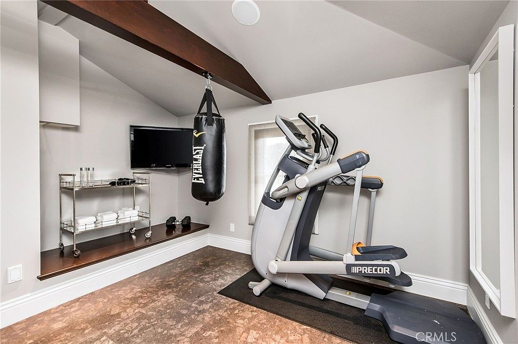 This home gym features a dedicated workout space equipped with a professional-grade elliptical machine and a hanging Everlast punching bag. A built-in dark wood shelf provides storage for towels and small weights, complemented by a rolling metal cart and a wall-mounted television. The room has a sloped ceiling with a dark wood beam and a unique, textured floor surface, creating a functional and private fitness area.