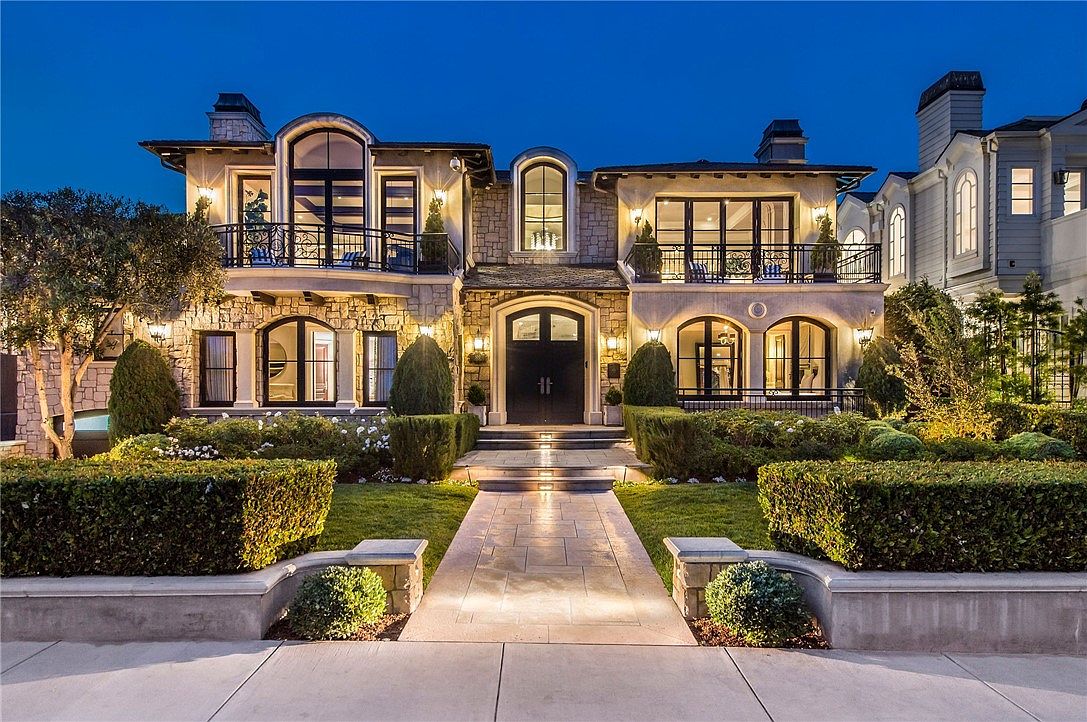 This stunning two-story luxury residence features a sophisticated stone facade with arched architectural details and symmetrical balconies. A manicured front garden with structured hedges leads to a grand, dark-toned double-door entrance, all illuminated by warm exterior lighting against a deep blue twilight sky. The property exudes an elegant, high-end aesthetic with a clean, inviting approach.