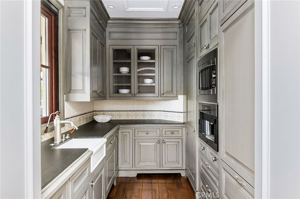 This compact, elegant kitchen area features custom grey cabinetry with glass-front upper cabinets and dark stone countertops. A classic farmhouse-style sink is positioned under a window, while a built-in microwave and wall oven are integrated into the cabinetry on the right. The space is finished with dark wood flooring and a decorative tile backsplash, creating a sophisticated and functional butler's pantry or secondary kitchen space.