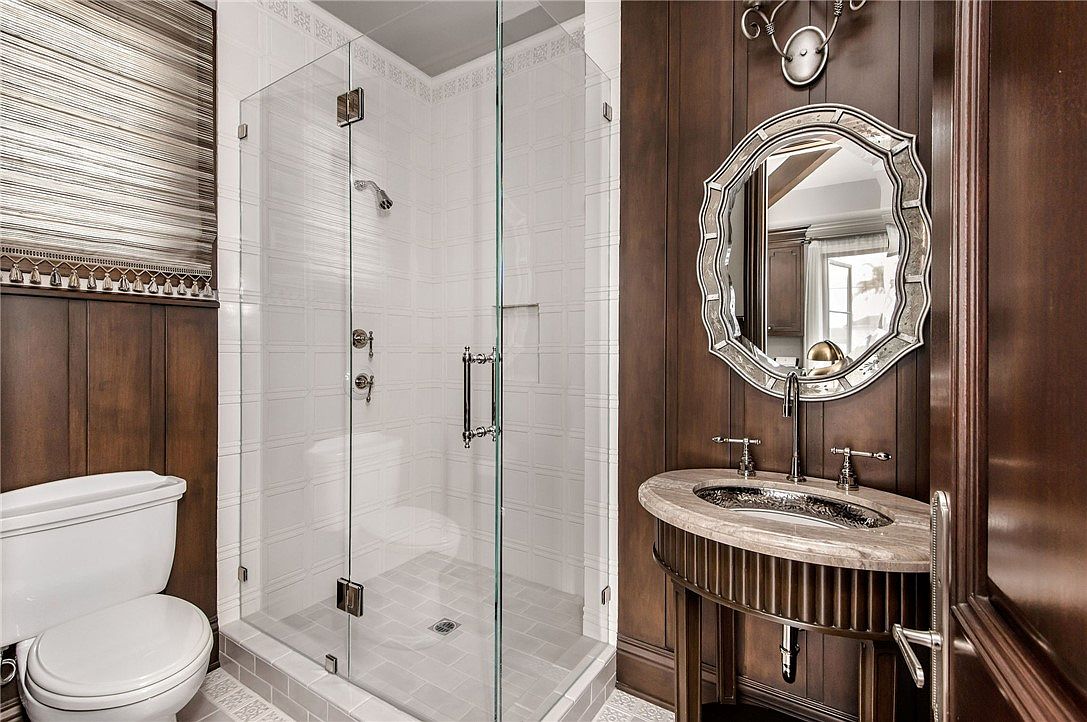 This elegant bathroom features dark wood-paneled walls that provide a sophisticated, traditional aesthetic. A glass-enclosed shower with white tiled walls and a gray tiled floor sits adjacent to a pedestal-style vanity with a stone countertop and a decorative, scalloped-edge mirror. The space is well-lit by a wall-mounted light fixture, creating a refined and polished atmosphere.