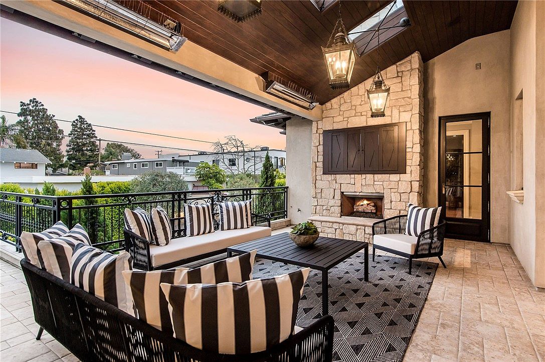 This covered outdoor living space features a stone-clad chimney breast with an integrated fireplace and television, creating a cozy focal point. The area is furnished with comfortable seating, including a sofa and armchairs with striped cushions, arranged on a patterned area rug. The space is open to the outdoors with a metal railing, offering a view of the neighborhood at sunset.