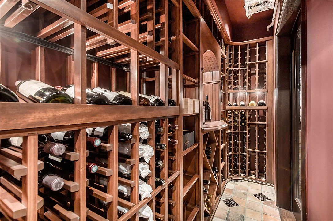 This sophisticated wine cellar features extensive custom wood racking designed to store a large collection of bottles horizontally. The space is finished with warm, dark-toned wood cabinetry and a patterned tile floor, creating an intimate and refined atmosphere. A small tasting counter is integrated into the shelving, and the room is illuminated by soft, ambient lighting that highlights the curated wine selection.