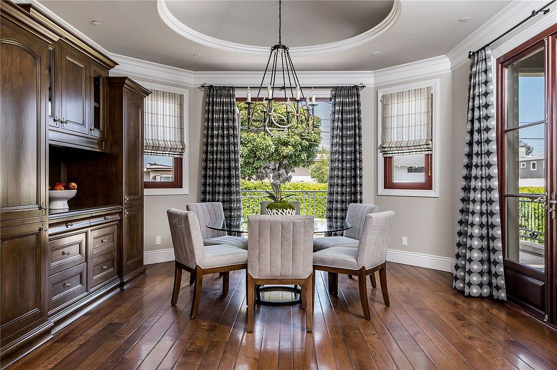 This elegant dining room features a circular tray ceiling with a central chandelier hanging above a glass-topped table surrounded by four upholstered chairs. To the left, a large, dark wood built-in cabinet provides ample storage, while the rear wall features large windows and glass doors draped in patterned curtains that open to a balcony. The room is finished with rich, dark wood flooring, creating a sophisticated and inviting atmosphere.