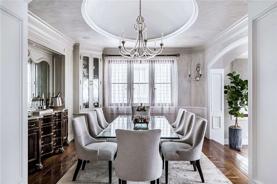 This elegant dining room features a glass-topped table surrounded by six upholstered chairs, set beneath a decorative chandelier centered in a circular ceiling medallion. To the left, a dark wood sideboard with a mirror and glassware provides a sophisticated serving area, while a corner cabinet with glass doors adds display space. The room is framed by tall windows draped in sheer curtains, and a large potted plant adds a touch of greenery to the corner, creating a refined and inviting atmosphere.