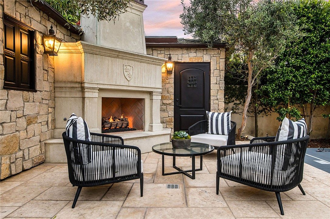This inviting outdoor patio features a prominent stone fireplace with a tall chimney breast, serving as the focal point of the space. The area is furnished with three modern, black-framed chairs with striped cushions arranged around a circular glass-top coffee table. The patio is paved with light-toned stone tiles and is framed by rustic stone walls, a dark wooden door, and lush greenery, creating a cozy and sophisticated atmosphere.