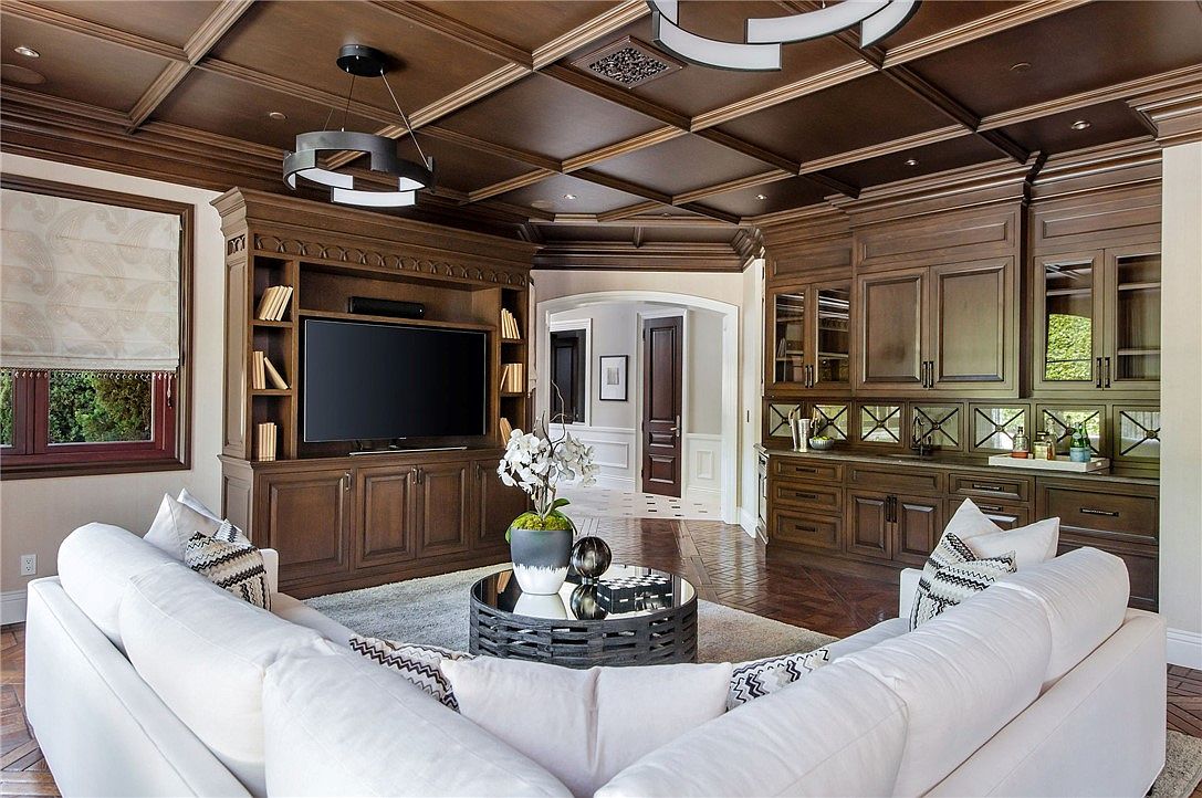 This sophisticated living room features a coffered dark wood ceiling and rich wood cabinetry that frames a large television and a built-in wet bar with mirrored backsplash. A plush, white sectional sofa provides ample seating around a circular, dark-toned coffee table, creating a cozy yet elegant atmosphere. The space is anchored by dark hardwood flooring and flows seamlessly into an adjacent hallway, showcasing a refined and cohesive interior design.