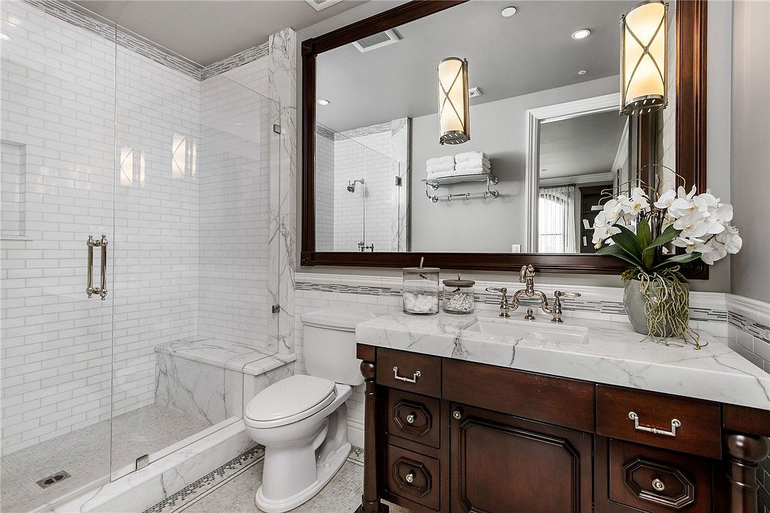 This elegant bathroom features a dark wood vanity topped with a light-colored stone counter and a large, framed mirror. To the left, a glass-enclosed shower is finished with white subway tile and a stone bench, while a white toilet sits between the shower and the vanity. The space is well-lit by wall-mounted sconces and recessed lighting, creating a sophisticated and polished atmosphere.