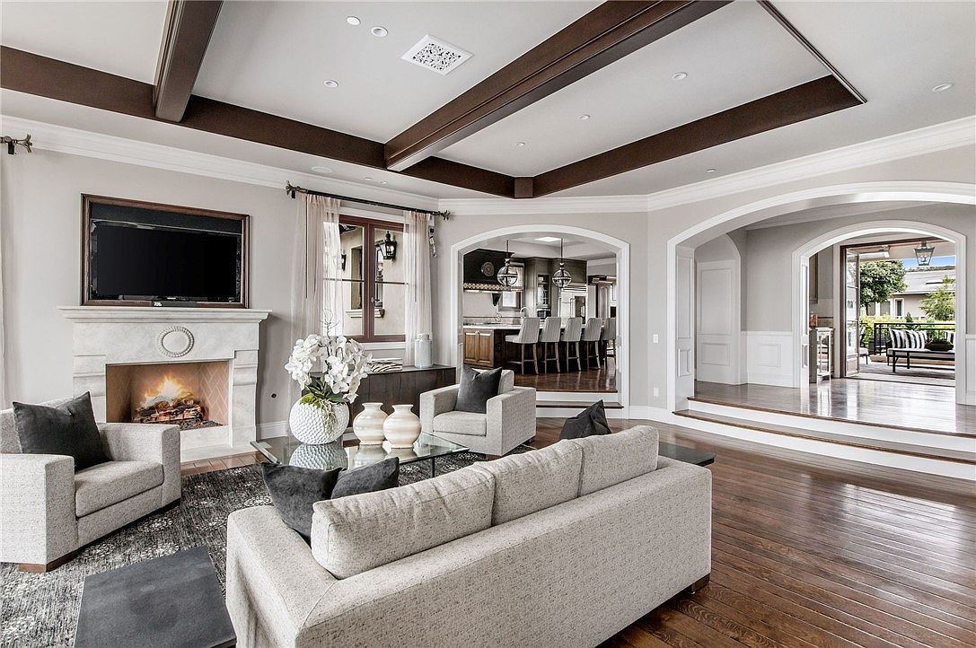 This elegant living room features a coffered ceiling with dark wood beams and a central fireplace with a stone surround. The space is furnished with a neutral-toned sofa and matching armchairs arranged on a textured area rug over dark wood flooring. An arched opening leads to a bright kitchen area, while another arched doorway provides a view of an outdoor patio space.