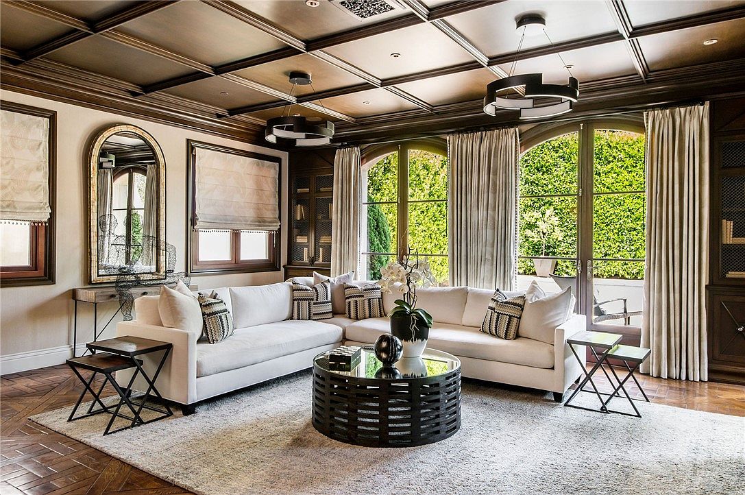 This sophisticated living room features a large, L-shaped white sectional sofa centered around a circular, slatted black coffee table. The space is defined by a coffered ceiling with dark wood beams and herringbone-patterned wood flooring, complemented by tall arched windows draped in neutral curtains. A decorative mirror and built-in shelving units add architectural interest, creating an elegant and inviting atmosphere.