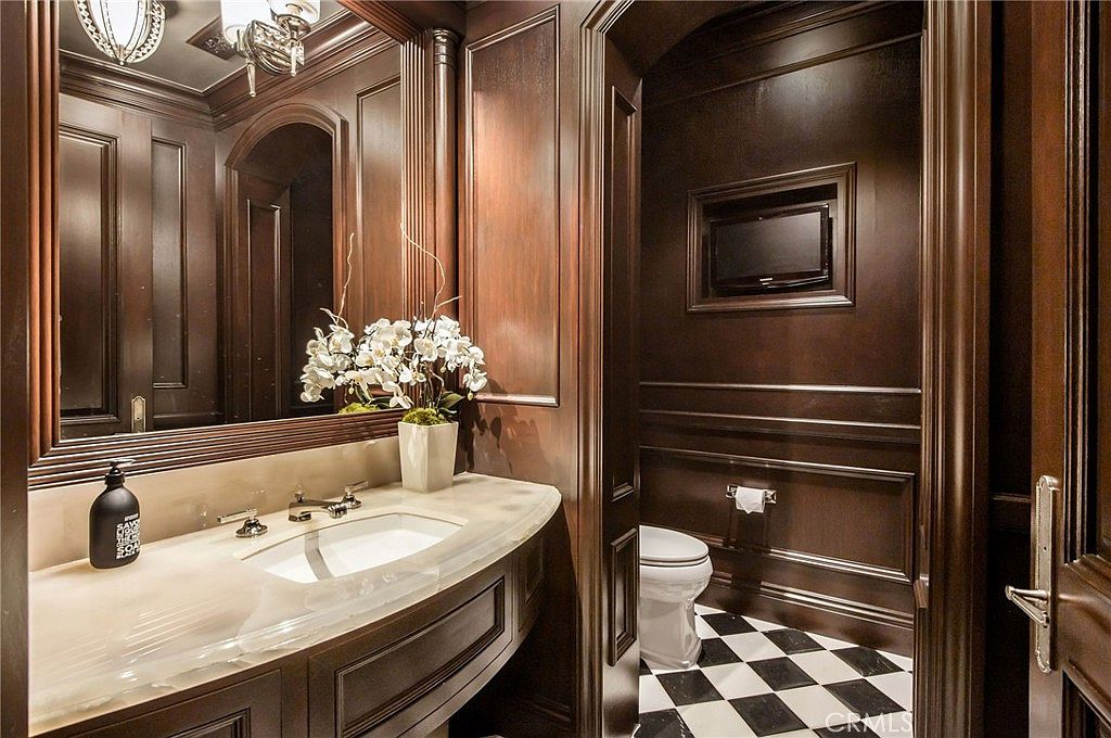 This elegant powder room features rich, dark wood paneling throughout, creating a sophisticated and moody atmosphere. The vanity is topped with a light-toned stone counter and includes a white undermount sink, while a large mirror reflects the space and a decorative floral arrangement. The floor is finished in a classic black-and-white checkered tile pattern, and a small television is mounted into the wall above the toilet area.