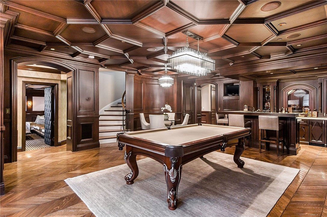 This sophisticated basement recreation area features rich, dark wood paneling and an intricate coffered ceiling, creating a luxurious, club-like atmosphere. The space is anchored by a central billiard table on a large area rug, complemented by a built-in bar area and a small dining nook near a staircase. The room is finished with high-end wood flooring laid in a herringbone pattern, providing a cohesive and elegant aesthetic throughout.