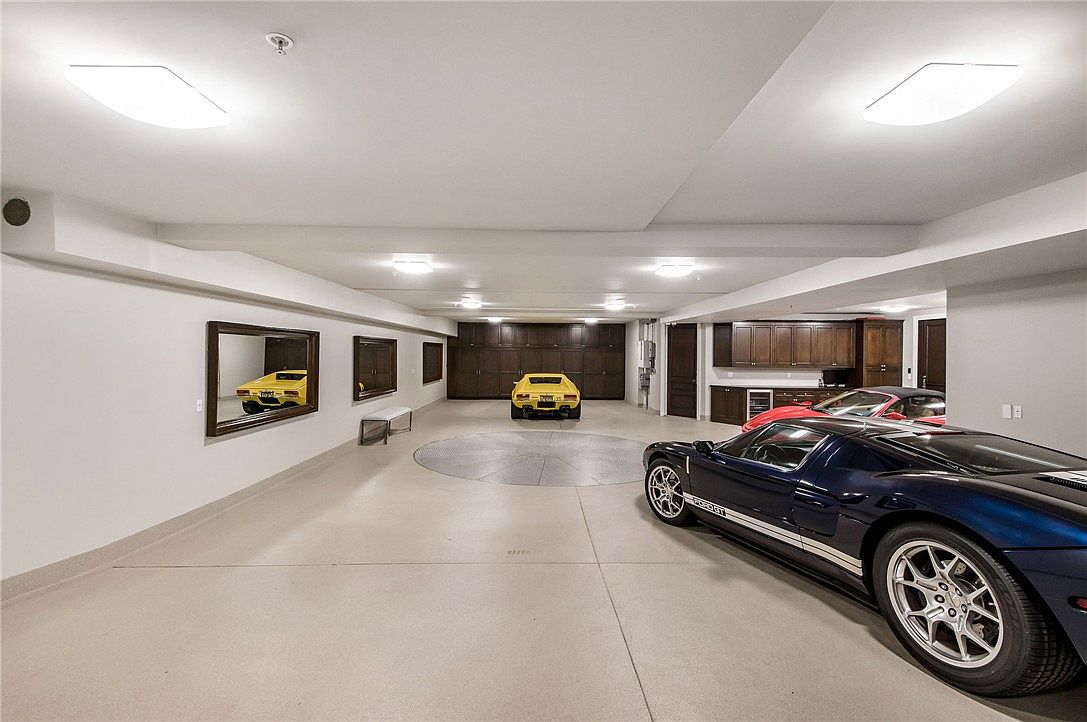 This expansive, high-end garage features a polished concrete floor and a central rotating turntable for vehicle display. The space is equipped with extensive dark wood cabinetry for storage and is illuminated by multiple recessed ceiling lights. A blue Ford GT sits in the foreground, while a yellow sports car is positioned on the turntable, creating a sophisticated showroom-like atmosphere.