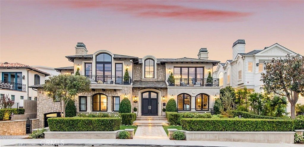 This grand, multi-story residence features a sophisticated stone facade with symmetrical architectural detailing and dark-framed windows. The front elevation includes two upper-level balconies with wrought-iron railings, a central arched window, and a formal entryway framed by manicured hedges and a stone-paved walkway. The scene is captured at dusk, with warm exterior lighting highlighting the home's elegant design against a soft, pastel-colored sky.