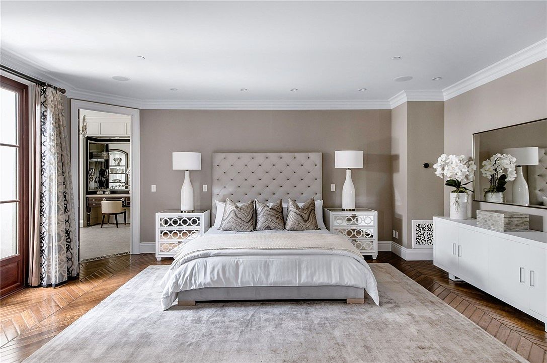 This elegant primary bedroom features a central tufted headboard flanked by mirrored nightstands and matching table lamps. A large area rug anchors the space, which includes a white console table topped with floral arrangements and a view into an adjacent dressing area. The room is finished with neutral wall tones and rich wood flooring, creating a sophisticated and serene atmosphere.