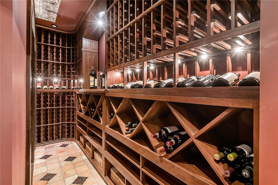This sophisticated wine cellar features extensive custom wood racking systems designed for both individual bottle storage and bulk display. The space is accented by warm, recessed lighting that highlights the collection, while the floor is finished with rustic, patterned terracotta-style tiles. The perspective is a wide-angle view from the entrance, showcasing the organized layout and the rich, dark wood tones that create an inviting and luxurious atmosphere.