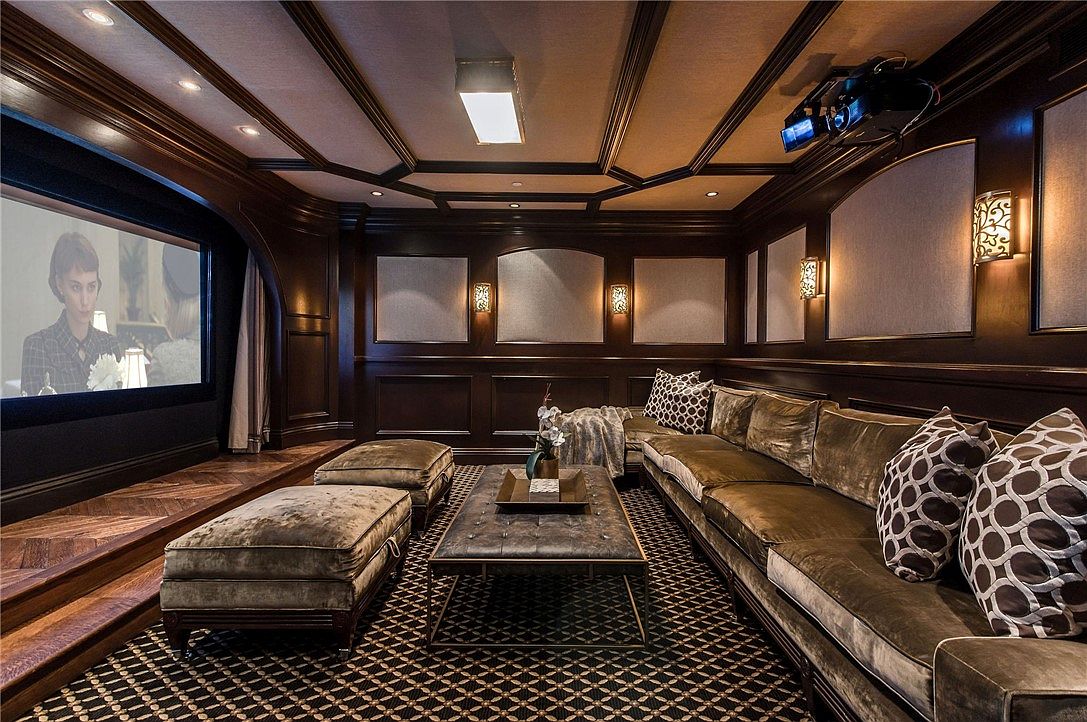 This luxurious home theater features dark wood paneling, coffered ceilings, and a large projection screen, creating an immersive cinematic environment. The room is furnished with a plush, velvet-upholstered sectional sofa, matching ottomans, and a central coffee table, all set atop a patterned carpet. Warm wall sconces provide ambient lighting, while a ceiling-mounted projector completes the professional media setup.