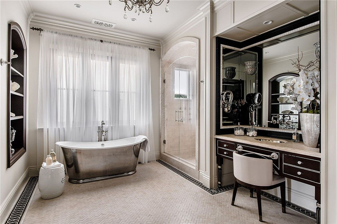 This luxurious primary bathroom features a metallic freestanding soaking tub positioned beneath large windows draped in sheer curtains. To the right, a dark-toned vanity with a stone countertop and a matching chair creates a sophisticated dressing area, while a glass-enclosed shower with stone tile walls sits between the tub and vanity. The room is finished with a herringbone-patterned tile floor and an elegant chandelier, conveying a high-end, spa-like atmosphere.