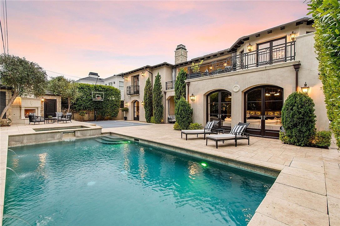 This luxurious backyard features a large, custom-shaped swimming pool with an integrated spa and water features. The expansive stone patio provides ample space for lounging, complemented by two sunbeds and an outdoor fireplace area in the background. The Mediterranean-style home showcases arched glass doors, a second-story balcony with wrought-iron railings, and manicured landscaping, creating an elegant and inviting atmosphere.