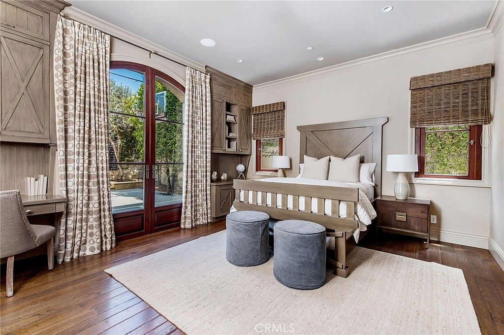 This elegant bedroom features a neutral color palette with warm wood tones and soft textures. A large bed with a wooden headboard serves as the focal point, flanked by matching nightstands and lamps, while two plush grey ottomans sit at the foot of the bed on a light-colored area rug. The room includes a built-in desk area and custom cabinetry, with large glass doors providing a view of the outdoor greenery.