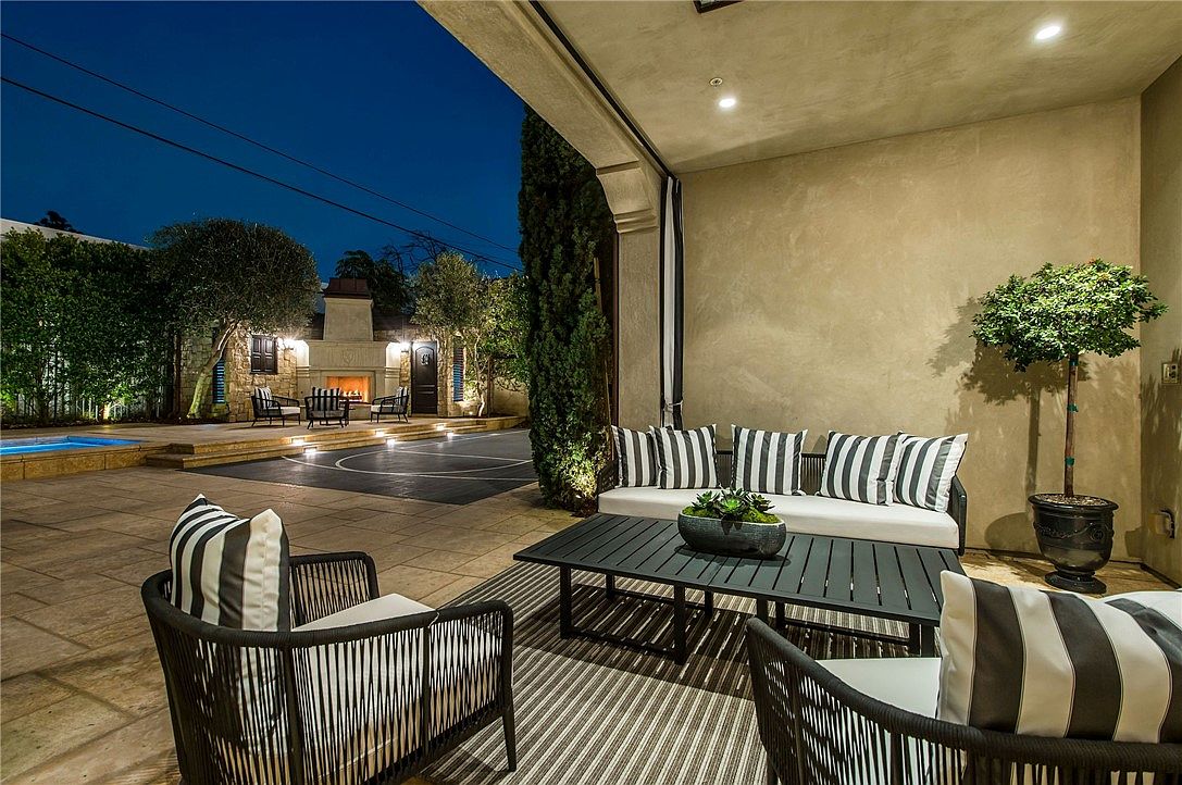This covered outdoor patio offers a sophisticated lounge area with modern black-and-white striped cushions and a low-profile coffee table. The space overlooks a beautifully landscaped backyard featuring a swimming pool, a stone-clad fireplace, and a paved recreation area. The perspective is from under the covered patio, looking out toward the illuminated evening landscape, creating an inviting and luxurious atmosphere for outdoor entertaining.