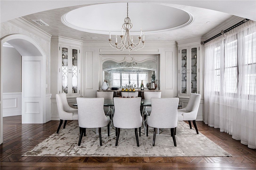 This elegant dining room features a central glass-topped table surrounded by six upholstered chairs, set beneath a decorative chandelier and a recessed circular ceiling detail. The room is anchored by a large area rug on dark wood flooring and includes built-in glass-front cabinetry flanking a central mirrored buffet area. The space is bathed in natural light from large windows draped in sheer curtains, creating a sophisticated and formal atmosphere.