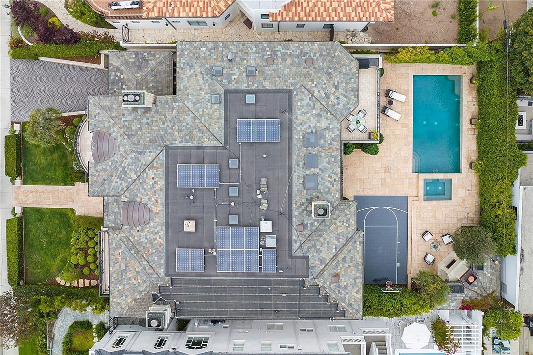 This high-angle aerial perspective captures a sprawling residential property featuring a complex, multi-faceted roofline with integrated solar panels. The grounds include a private swimming pool, a dedicated basketball court, and meticulously landscaped green spaces bordered by tall hedges. The layout emphasizes a luxurious outdoor living experience, showcasing a seamless blend of recreational amenities and structured architectural design.