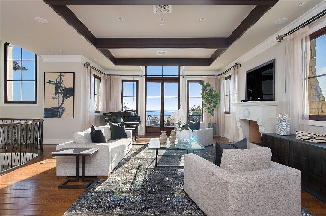 This elegant living room features a coffered ceiling with dark wood beams and a central seating area arranged around a glass-topped coffee table. A stone fireplace with a television mounted above serves as a focal point, while large windows and glass doors provide abundant natural light and a view of the ocean. The space is anchored by a patterned area rug over hardwood floors, complemented by a grand piano positioned near the rear windows.