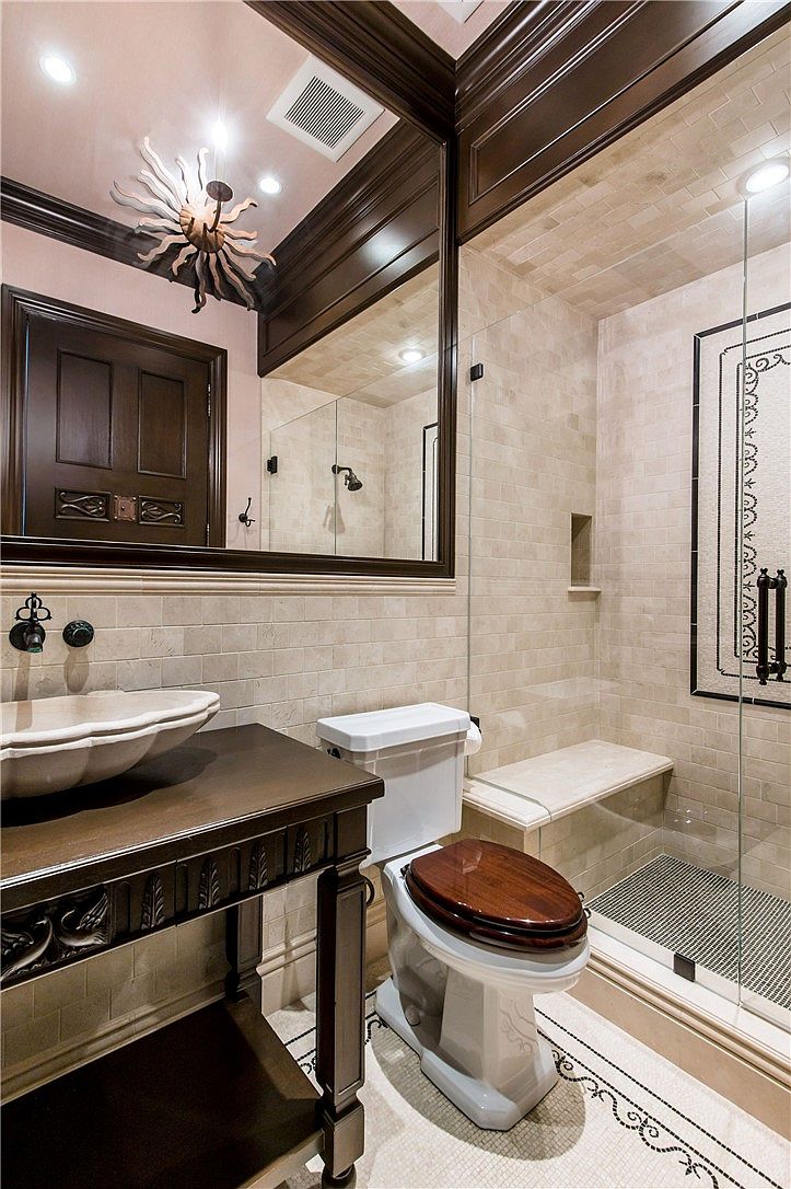 This elegant bathroom features a unique shell-shaped vessel sink set atop a dark wood vanity with intricate carved details. A glass-enclosed shower with light-toned wall tiles and a built-in bench sits adjacent to a toilet with a dark wood seat. The space is accented by a large mirror with a dark frame, a decorative sunburst-style light fixture, and a patterned tile border on the floor.