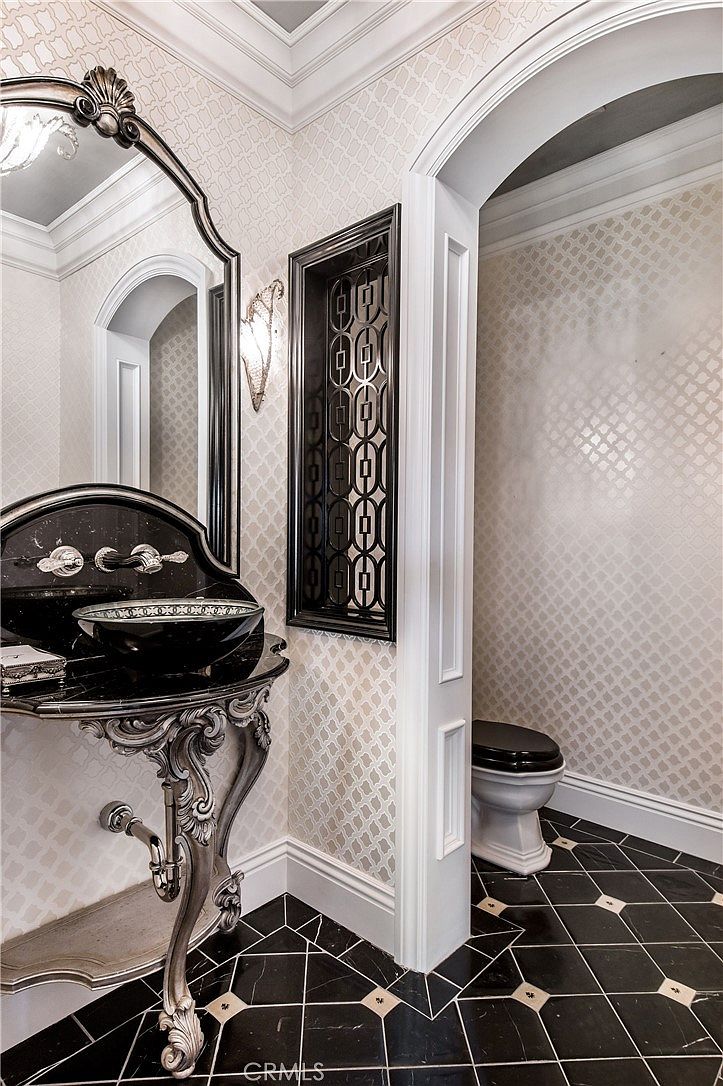 This elegant powder room features a sophisticated black and white color palette with ornate, traditional design elements. A pedestal-style vanity with a dark stone basin and metallic legs sits beneath a large, arched mirror, while a decorative black metal wall screen adds a dramatic architectural touch. The space is finished with patterned wallpaper and classic black-and-white tiled flooring, creating a luxurious and refined atmosphere.