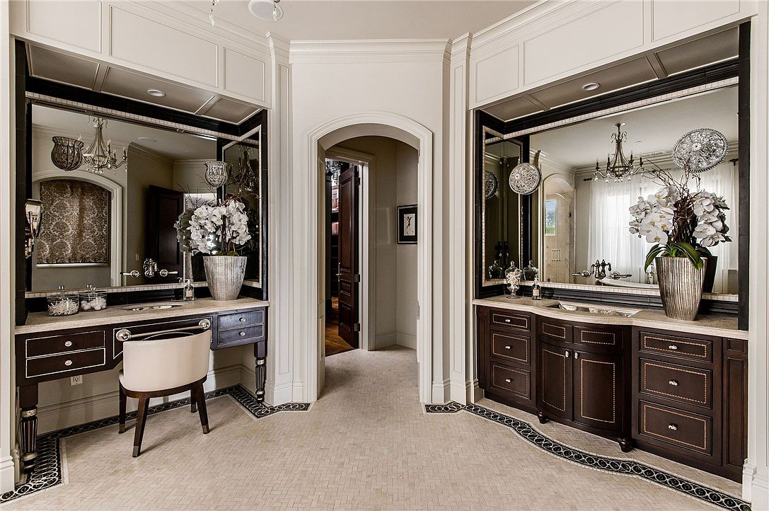 This elegant primary bathroom features a symmetrical layout with dual dark wood vanities flanking a central arched doorway. The space is adorned with large mirrors, decorative floral arrangements, and intricate patterned tile borders on the floor. The neutral wall paneling and sophisticated lighting fixtures create a luxurious and refined atmosphere.