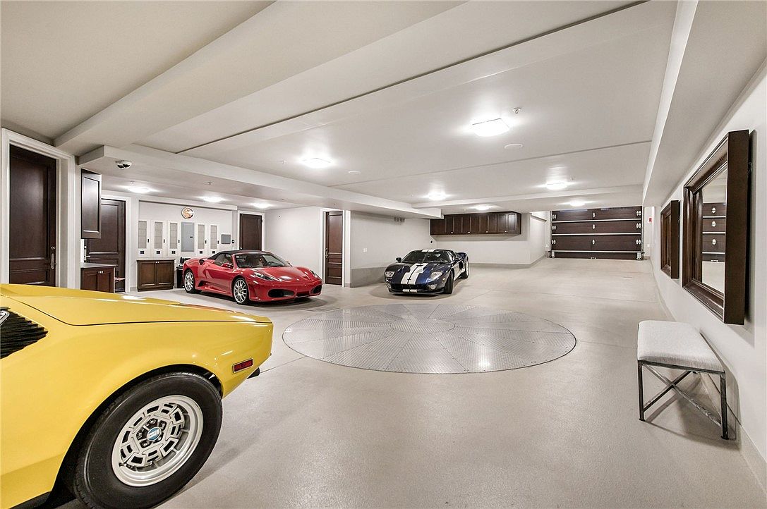 This expansive, high-end luxury garage features a central circular car turntable surrounded by a polished concrete floor. The space is illuminated by recessed ceiling lights and houses three high-performance sports cars, including a yellow model in the foreground, a red Ferrari, and a blue Ford GT. Dark wood cabinetry and doors provide a sophisticated contrast to the neutral walls, creating a clean, gallery-like atmosphere for vehicle storage.