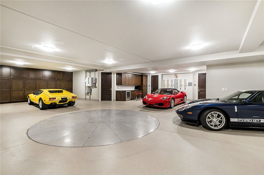 This expansive, high-end garage features a central circular car turntable surrounded by a polished concrete floor. The space is equipped with extensive dark wood cabinetry for storage, a wet bar area, and recessed ceiling lighting, housing three luxury vehicles. The perspective is a wide-angle shot capturing the entire showroom-style layout, emphasizing the professional-grade organization and premium aesthetic.