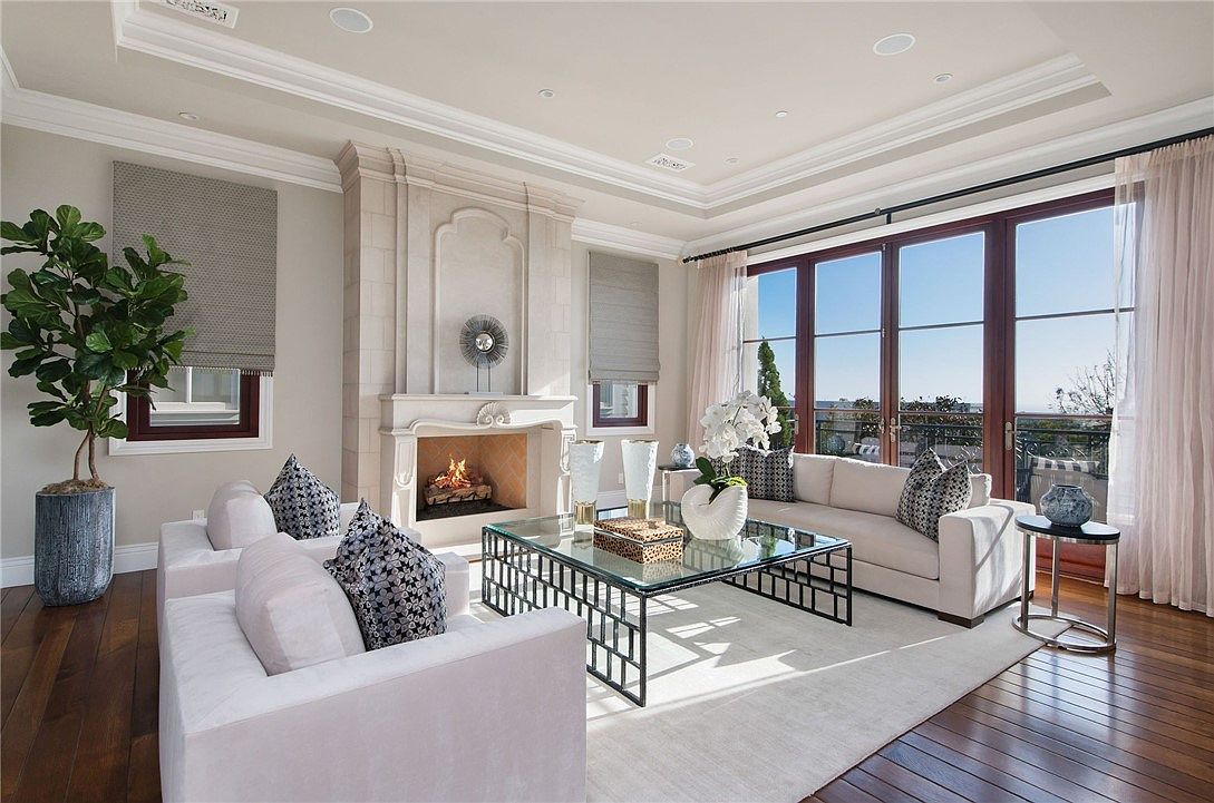 This elegant living room features a sophisticated neutral color palette, centered around a prominent fireplace with a tall, light-toned stone surround. The space is furnished with plush white sofas and a geometric-based glass coffee table, all set atop a large area rug on dark hardwood floors. Large windows and glass doors provide abundant natural light and a view of the outdoor balcony, creating an airy and luxurious atmosphere.