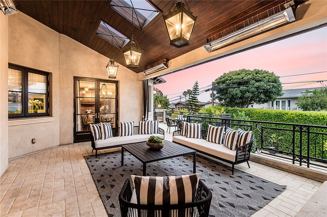 This covered outdoor patio features a sophisticated lounge area with two sofas and a central coffee table arranged on a geometric patterned rug. The space is defined by a dark wood-paneled ceiling with skylights and hanging lanterns, while a wrought-iron railing overlooks a lush green hedge and neighborhood trees at sunset. The open-air design creates a seamless transition between indoor and outdoor living, enhanced by mounted heaters for year-round comfort.