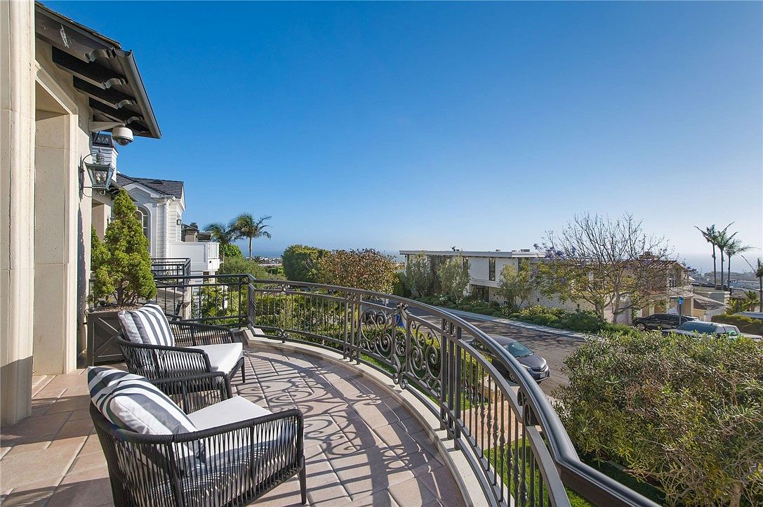 This elevated balcony offers a sweeping view of a quiet suburban street and the distant coastline under a clear blue sky. The space is furnished with two modern, dark-toned chairs featuring striped cushions, set against an ornate wrought-iron railing. The perspective captures the elegant architectural detail of the home's exterior while showcasing the scenic, open-air vantage point.