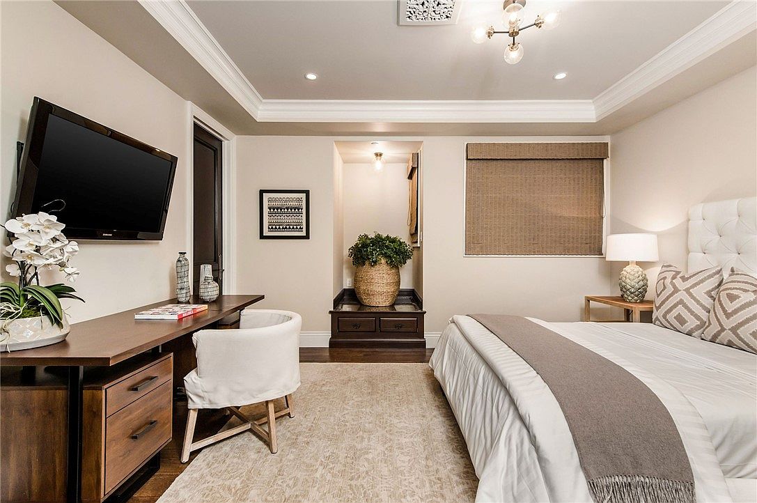 This elegant primary bedroom features a neutral color palette, a plush tufted headboard, and a dedicated workspace with a desk and chair. A recessed alcove displays a large potted plant atop a dark wood cabinet, adding a touch of nature to the sophisticated space. The room is illuminated by a modern chandelier and recessed lighting, creating a warm and inviting atmosphere.