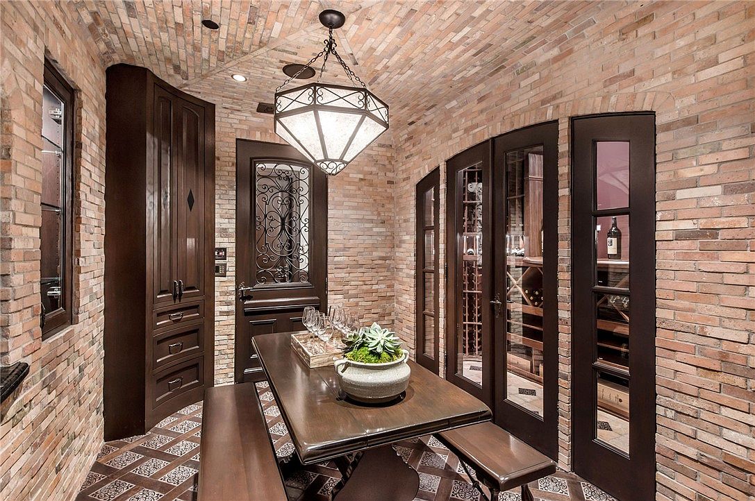 This sophisticated wine cellar features a barrel-vaulted ceiling and walls constructed from rustic, textured brick. The space is furnished with a dark wood table and matching benches, illuminated by a large, geometric pendant light fixture. Glass-paned doors reveal organized wine storage racks, creating an elegant and intimate atmosphere for tasting and entertaining.