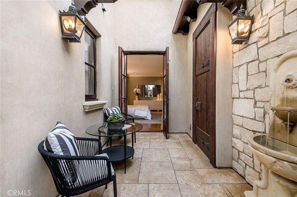This intimate, Mediterranean-style courtyard features a stone-tiled floor and a wall-mounted stone fountain, creating a serene outdoor retreat. A pair of black wicker chairs with striped cushions and a glass-topped bistro table are arranged near an open doorway that leads into a bedroom. The space is framed by warm-toned stucco walls, decorative lanterns, and a heavy wooden door, offering a sophisticated and private transition between indoor and outdoor living.